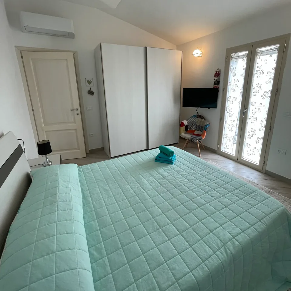 Camera matrimoniale con copriletto verde acqua, soffitto inclinato e portafinestra